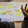Illustration d'une séance de travail collaboratif et de co-design (une main devant un mur de post-it)