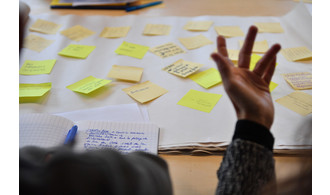Illustration d'une séance de travail collaboratif et de co-design (une main devant un mur de post-it)